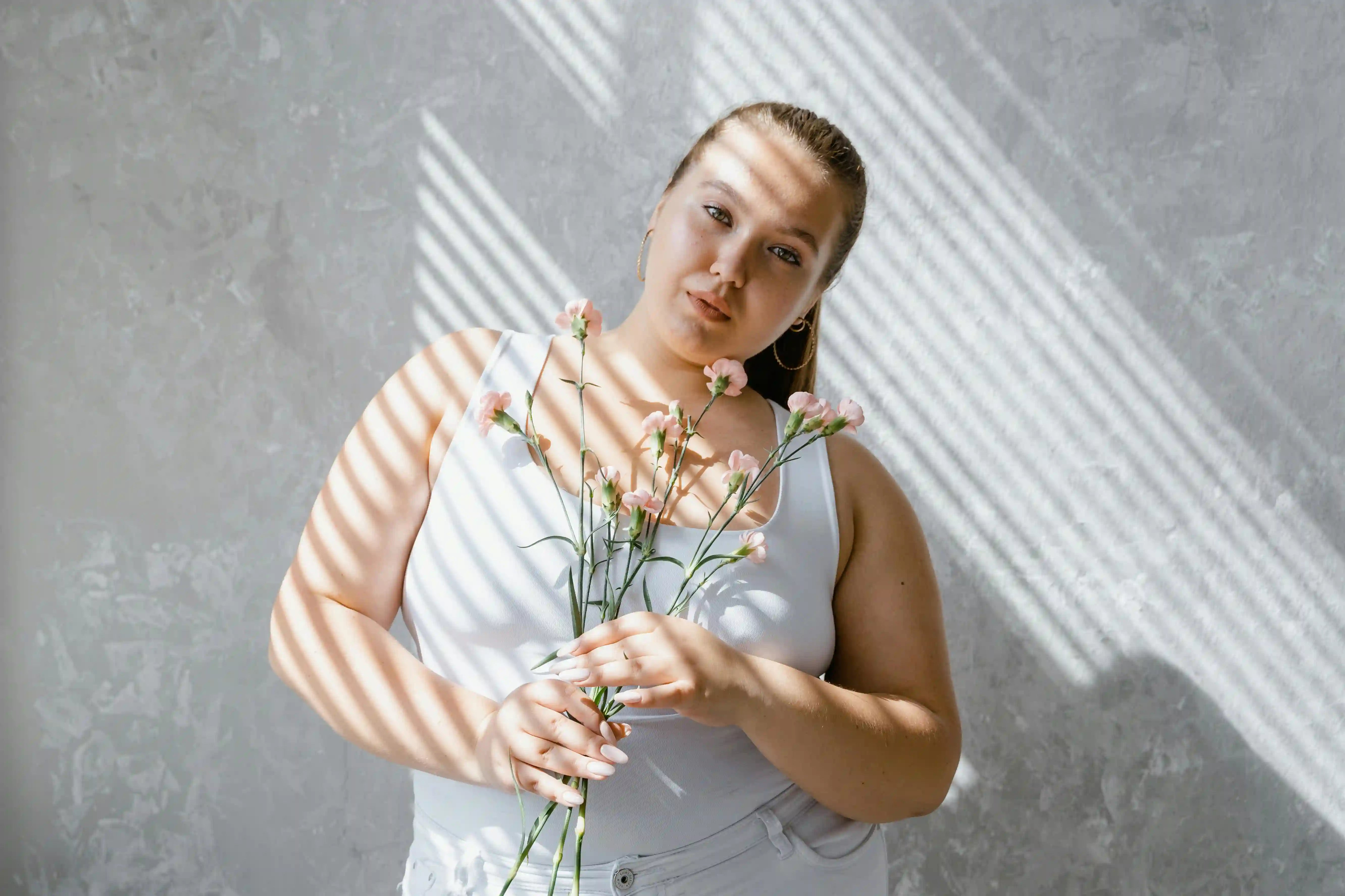 Woman in a white tank holding pink flowers, striped sunlight across her face and arms against a textured gray wall